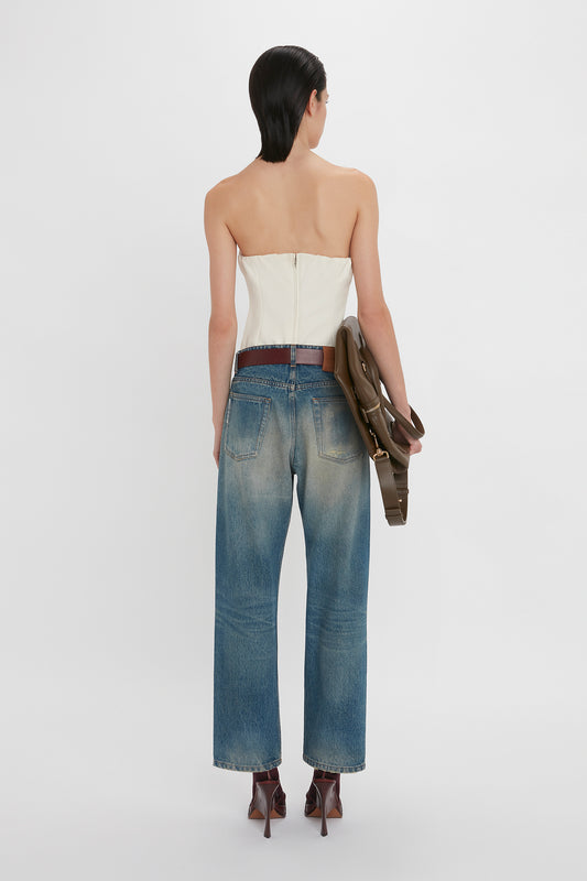 A woman wearing a strapless Antique White corset top by Victoria Beckham and blue jeans, seen from the back, holding a brown handbag.