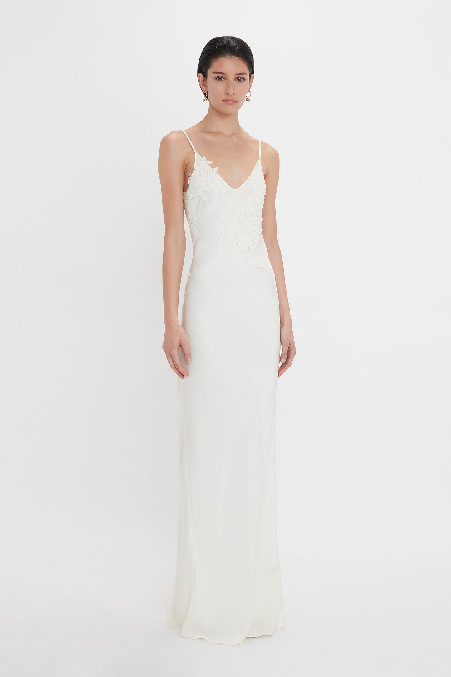 A woman in a white sleeveless lace-top wedding gown and Victoria Beckham's Exclusive Camellia Flower Hoop Earrings In Gold standing against a plain white backdrop.