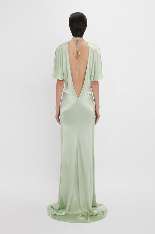 A woman in a Victoria Beckham exclusive floor-length jade satin gown with an open back and gathered skirt, standing against a white background.
