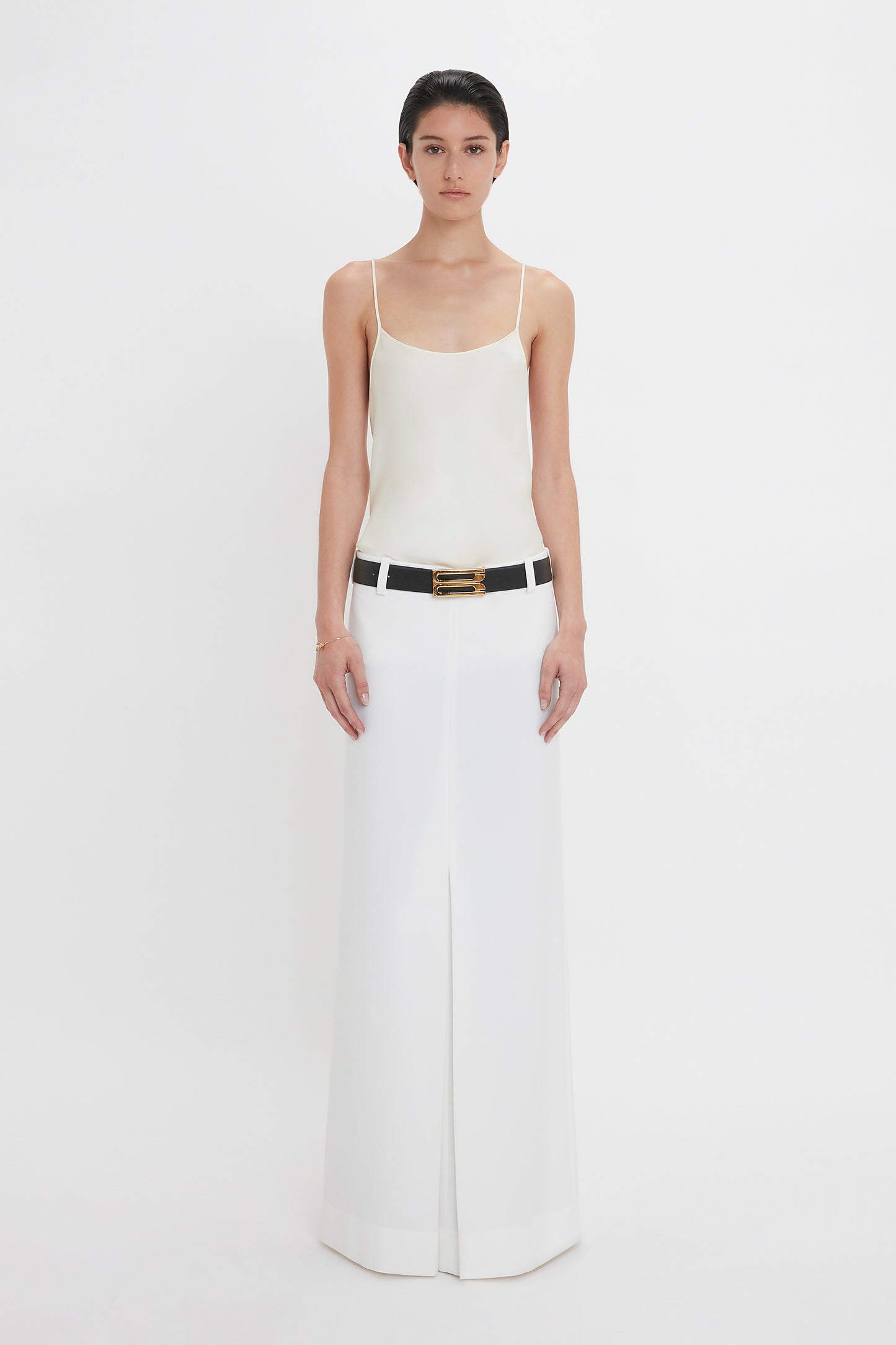 A person stands against a white background, wearing an Exclusive Cami Top In Ivory by Victoria Beckham and a long white skirt with a black belt featuring a gold buckle.