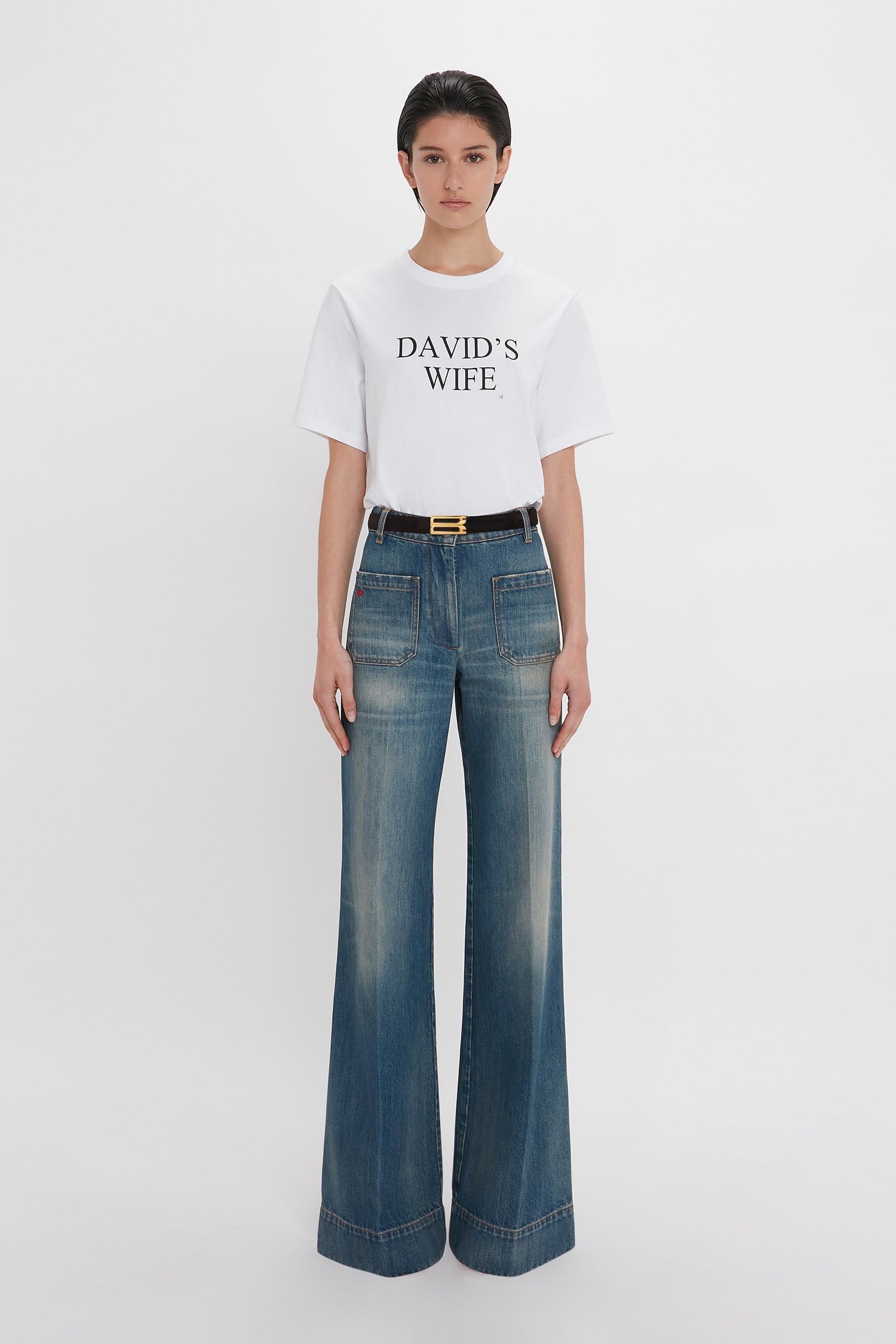 A woman wearing a white t-shirt with "david's wife" text and high-waisted Alina Jean In Indigrey Wash jeans by Victoria Beckham, accessorized with a black belt with a gold buckle, standing against a
