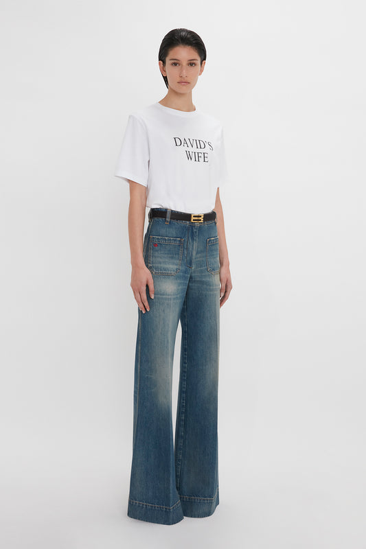 A woman wearing a white "Victoria Beckham" slogan T-shirt with the text "David's wife" and blue flared jeans, standing against a plain white background.
