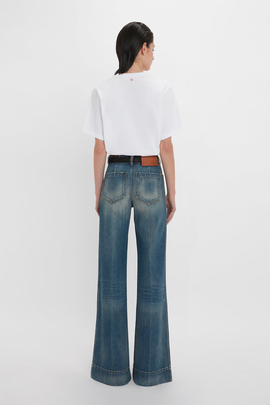 Woman in a white t-shirt and blue Victoria Beckham Alina Jean In Indigrey Wash flared jeans standing against a white background, viewed from the back.