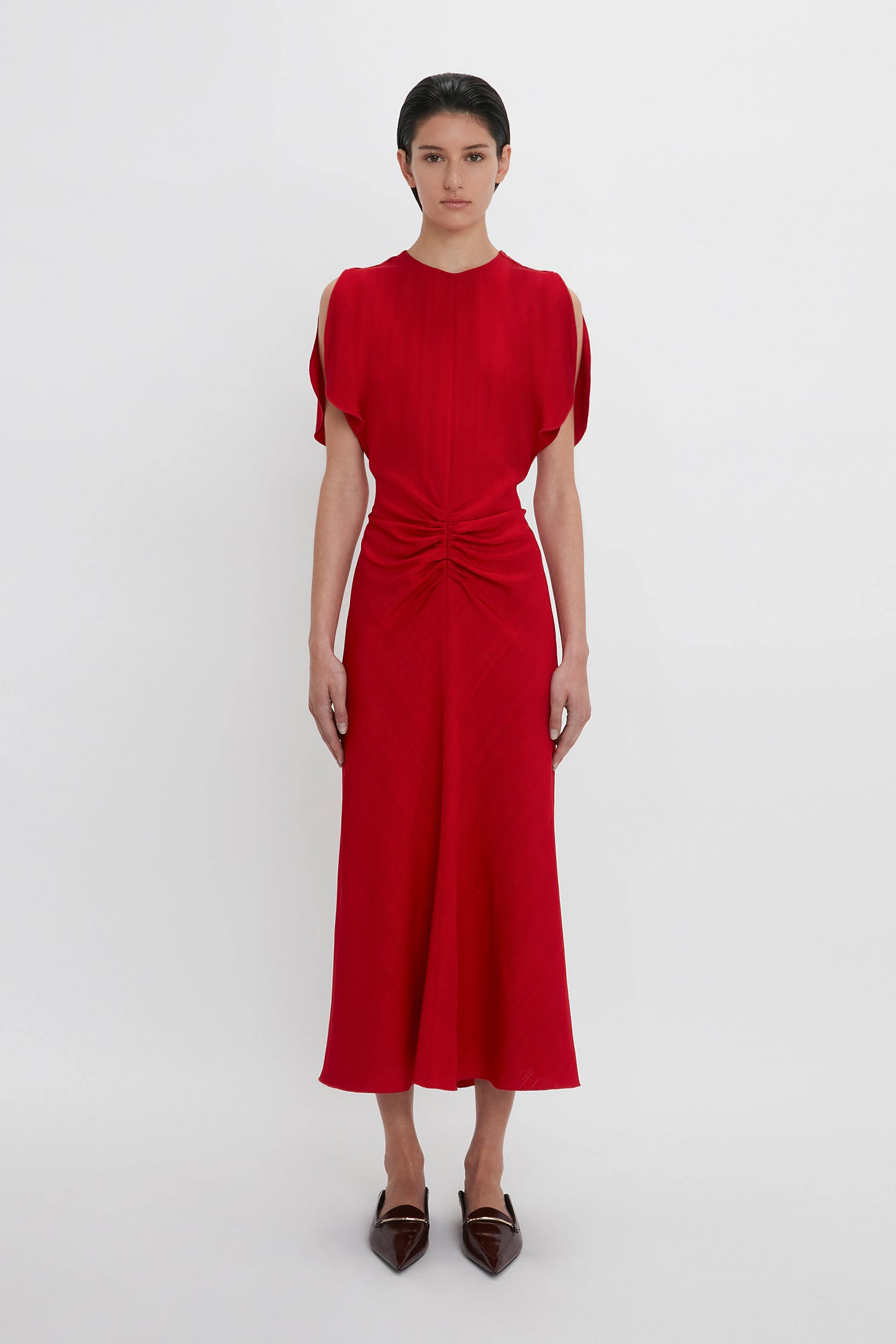 Woman in a red Victoria Beckham Exclusive Gathered V-Neck Midi Dress In Carmine with a twisted front detail, standing against a white background.