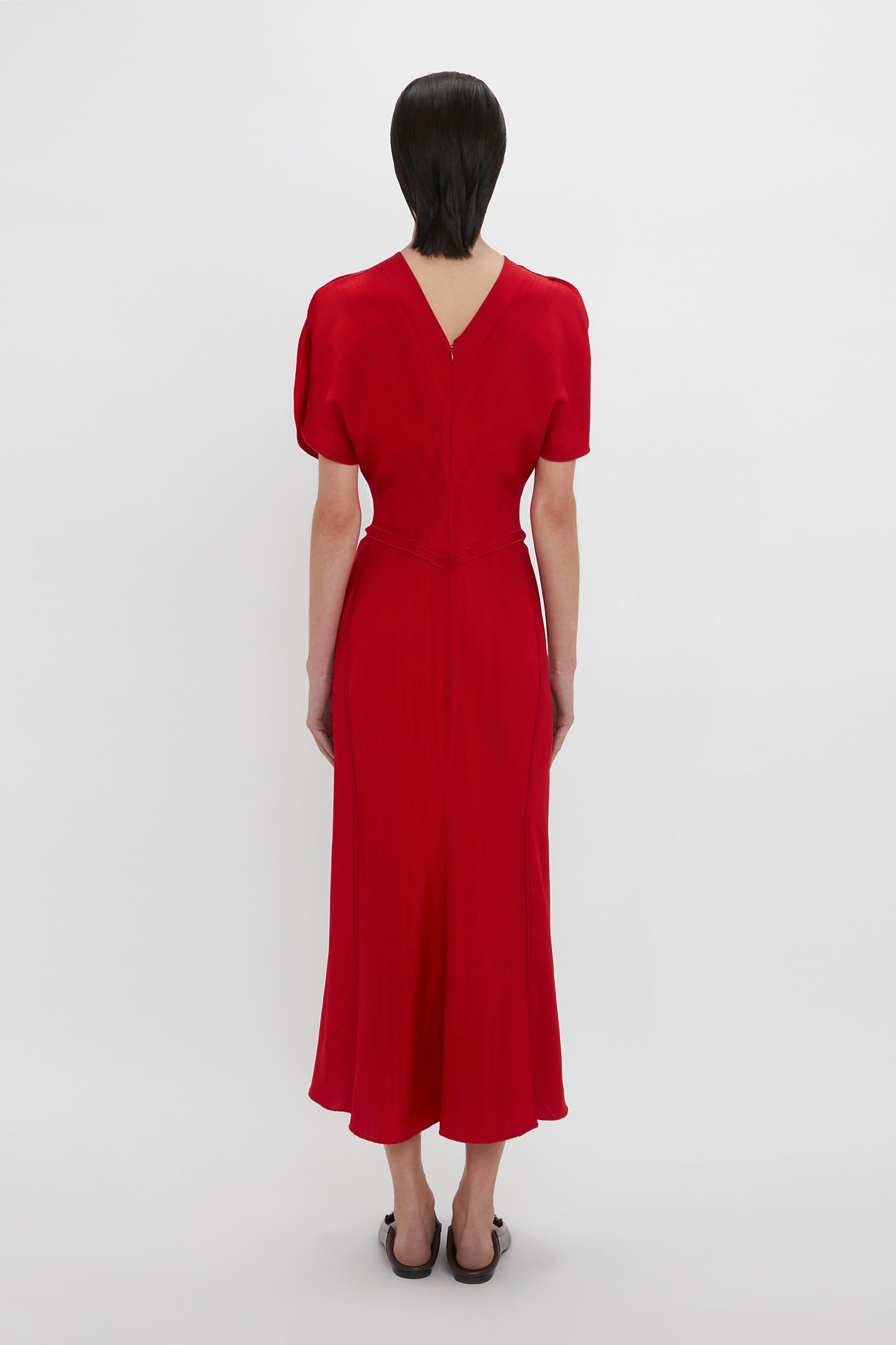 A woman stands with her back to the camera, wearing a Victoria Beckham red midi dress with short sleeves and a v-shaped neckline at the back. The dress features an elegant fit-and-flare silhouette.
