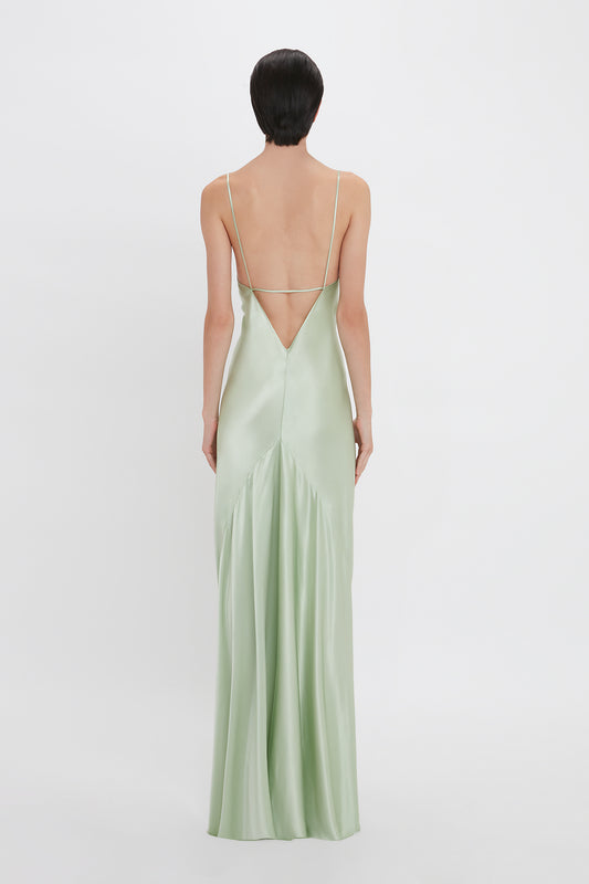 A woman with short black hair, viewed from behind, wears a Victoria Beckham pale green Exclusive Low Back Cami Floor-Length Dress In Jade with crisscross straps.