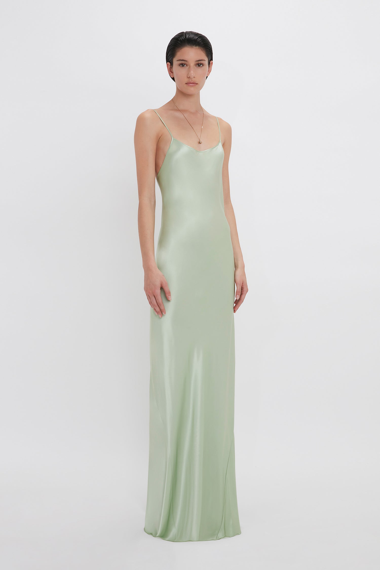 A woman stands against a white background wearing a long, light green Exclusive Low Back Cami Floor-Length Dress in Jade by Victoria Beckham with thin straps and a delicate camellia flower necklace.