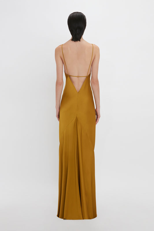 A woman stands with her back to the camera, wearing a Victoria Beckham mustard yellow slip dress with a low-cut, strappy back and a floor-length skirt.