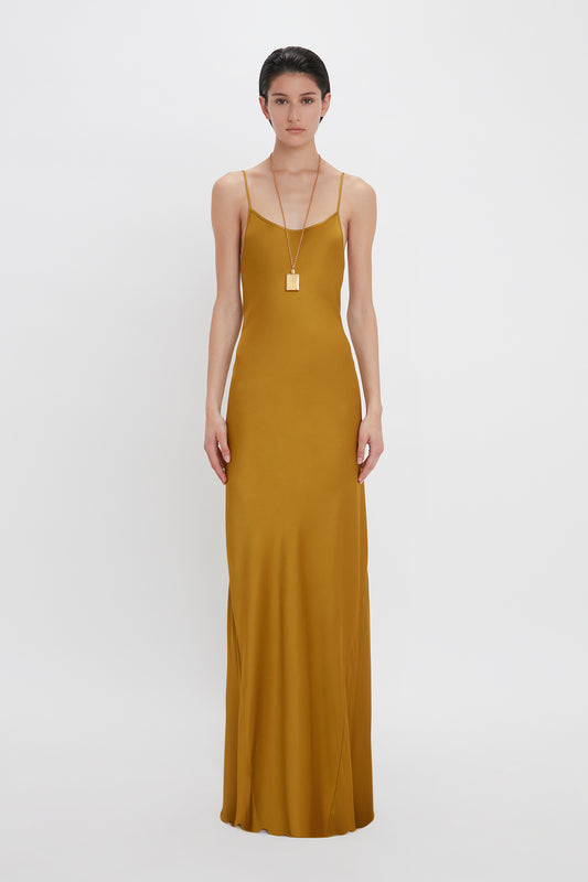 A woman in a Victoria Beckham Low Back Cami Floor-Length Dress In Harvest Gold and a long necklace, standing against a white background.