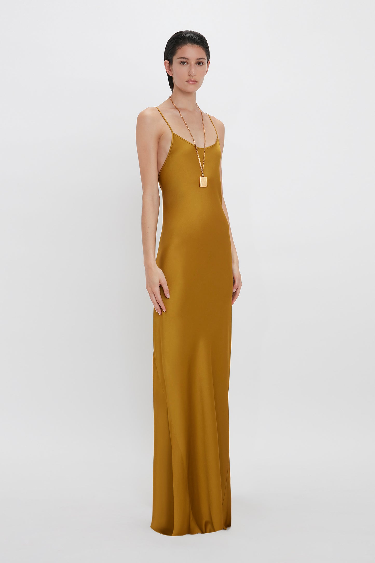 A woman in a long, mustard-colored Victoria Beckham Low Back Cami Floor-Length Dress In Harvest Gold with thin straps, standing against a white background. She wears a long gold necklace.