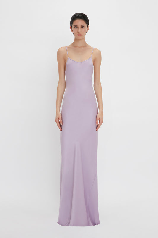 A woman stands against a white background, wearing a long, lilac floor-length Low Back Cami Dress In Petunia from Victoria Beckham with thin straps.