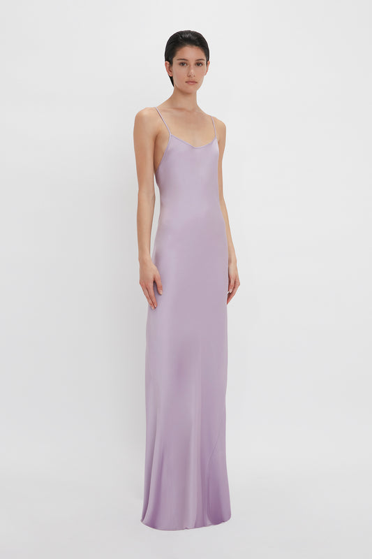 A woman in a long, lilac Low Back Cami Floor-Length Dress In Petunia by Victoria Beckham stands against a plain white background, looking directly at the camera.