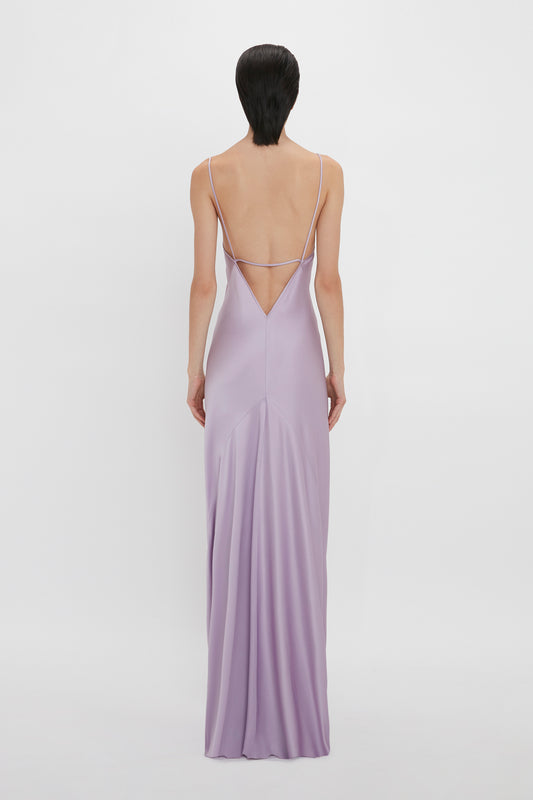 A woman stands with her back to the camera, wearing a long, lavender floor-length Low Back Cami Dress In Petunia with thin straps and a low back by Victoria Beckham.