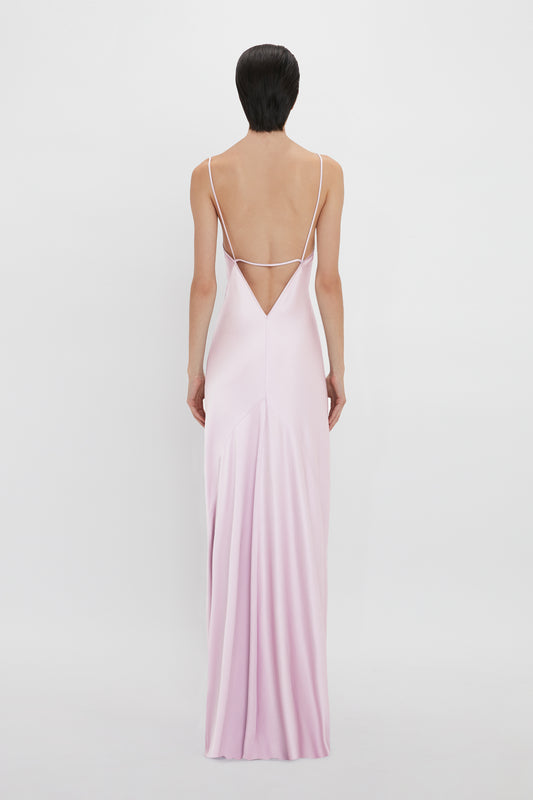 A woman with short dark hair stands with her back facing the camera, wearing a pink, floor-length slip dress with thin straps and a low-cut back - Victoria Beckham's Low Back Cami Floor-Length Dress In Rosa.