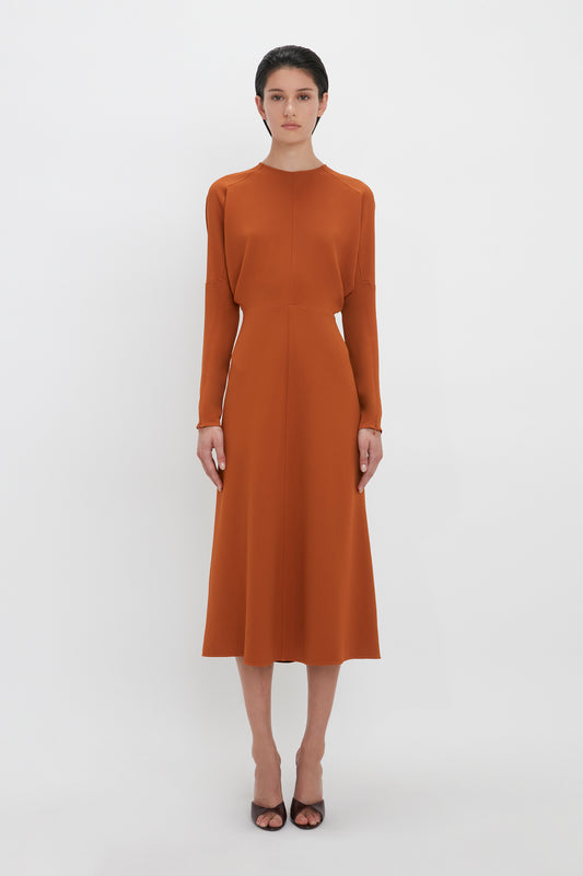 A woman in a sleek, long-sleeve Victoria Beckham Dolman Midi Dress In Russet stands against a plain white background, facing the camera with a neutral expression.