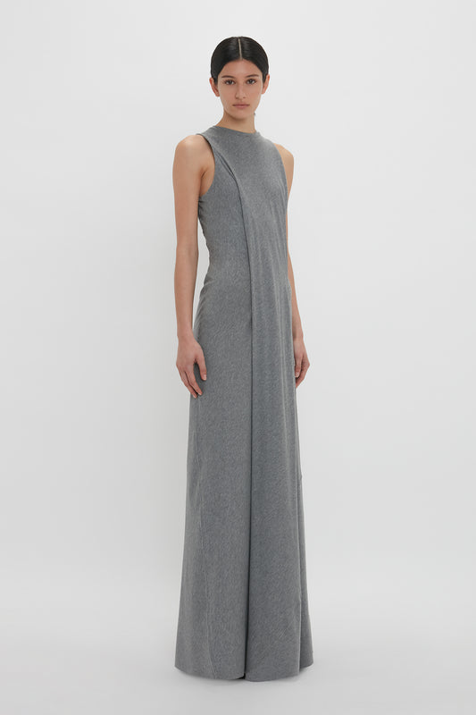 A woman stands in a studio, wearing a long, gray Victoria Beckham maxi dress with a high neckline and an elegant drape. She looks directly at the viewer, with a neutral expression.