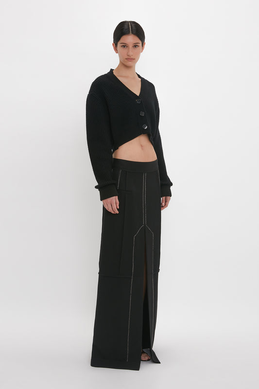 Woman standing in a studio wearing a black ribbed knit Victoria Beckham cropped v-neck cardigan and high-waisted trousers with unique stitching.