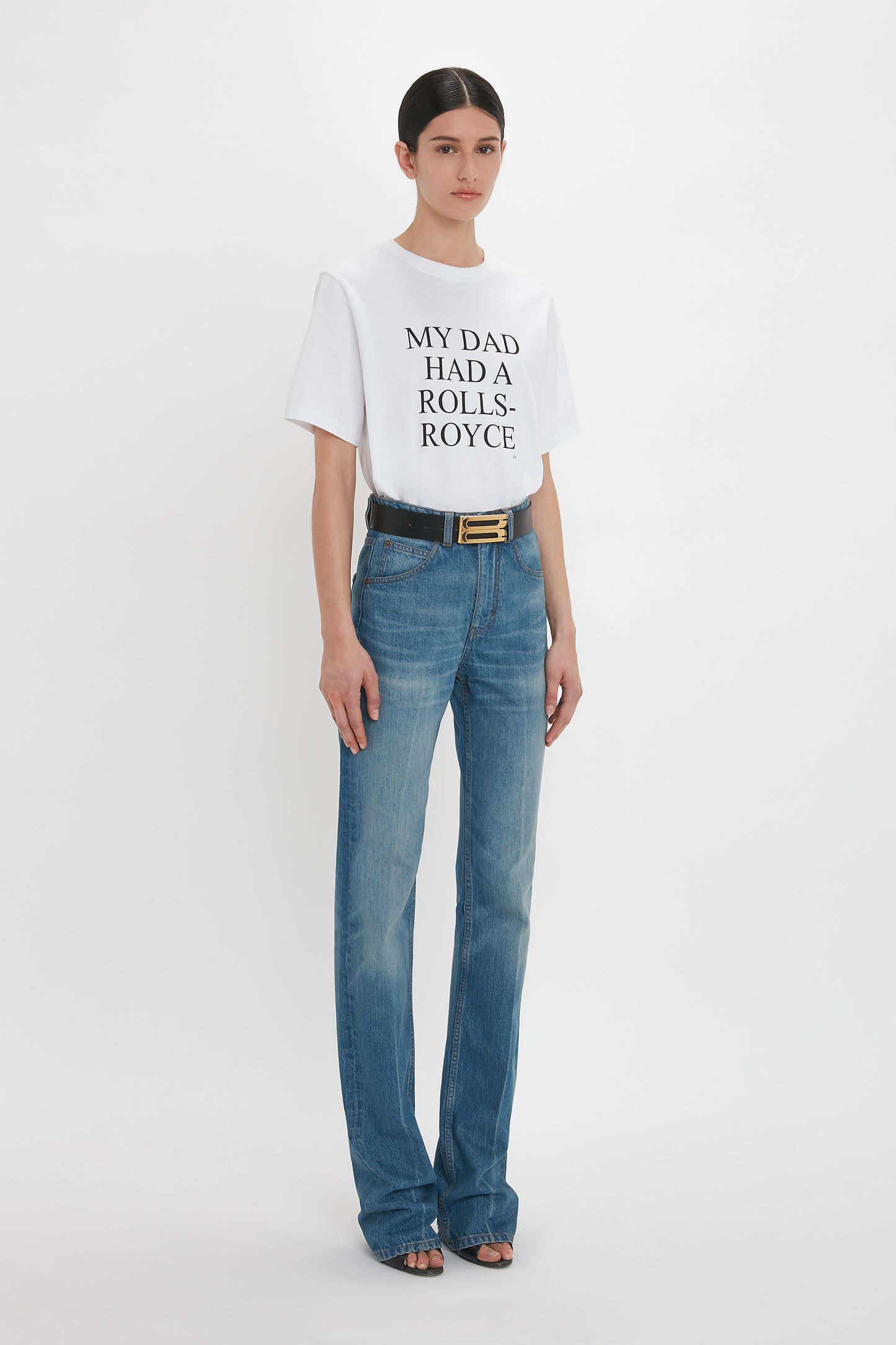 A woman wearing Victoria Beckham's Julia Jean In Broken Vintage Wash high waist jeans and a white t-shirt that reads "my dad had a rolls-royce" standing against a white background.