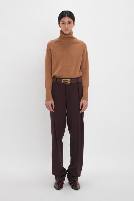A woman stands against a plain background, dressed in a Victoria Beckham camel luxury knitwear turtleneck sweater and dark purple trousers with a brown belt and burgundy shoes.