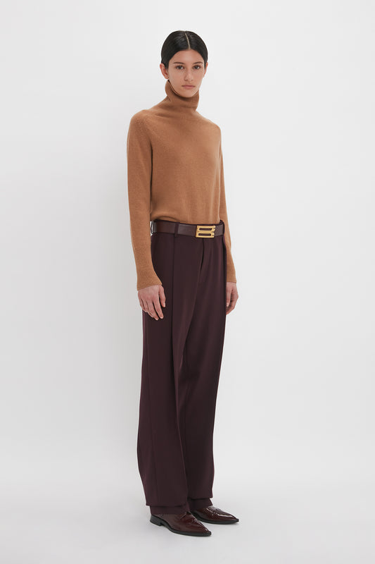 A woman stands against a white background, wearing a Victoria Beckham luxury knitwear lambswool polo neck jumper in tobacco, brown belt, dark trousers, and brown shoes.