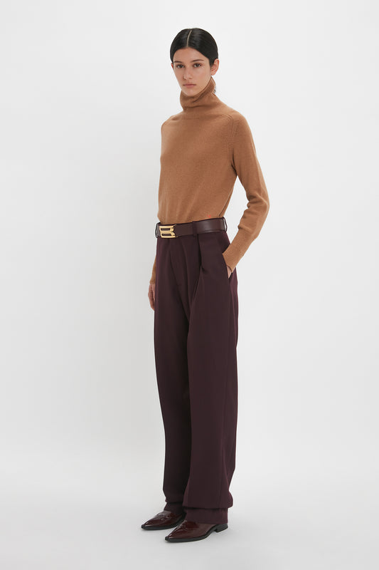 A woman standing in a studio, dressed in a brown lambswool Victoria Beckham polo neck sweater and dark brown high-waisted trousers with a belt, paired with dark brown shoes.