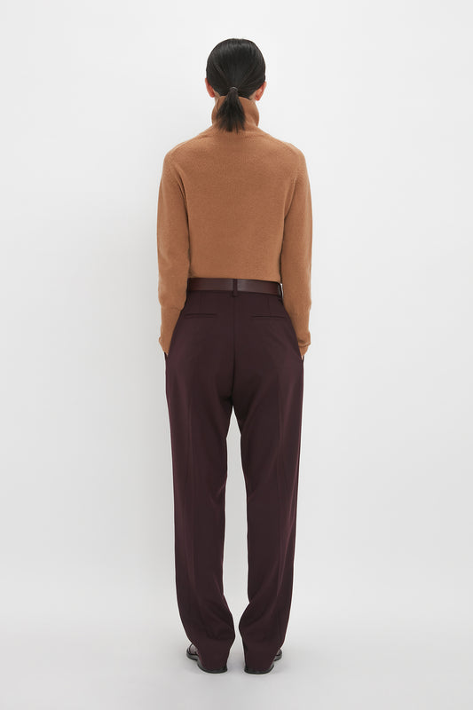 Rear view of a woman in a Victoria Beckham luxury knitwear camel-colored lambswool polo neck jumper and dark maroon trousers, standing against a white background.