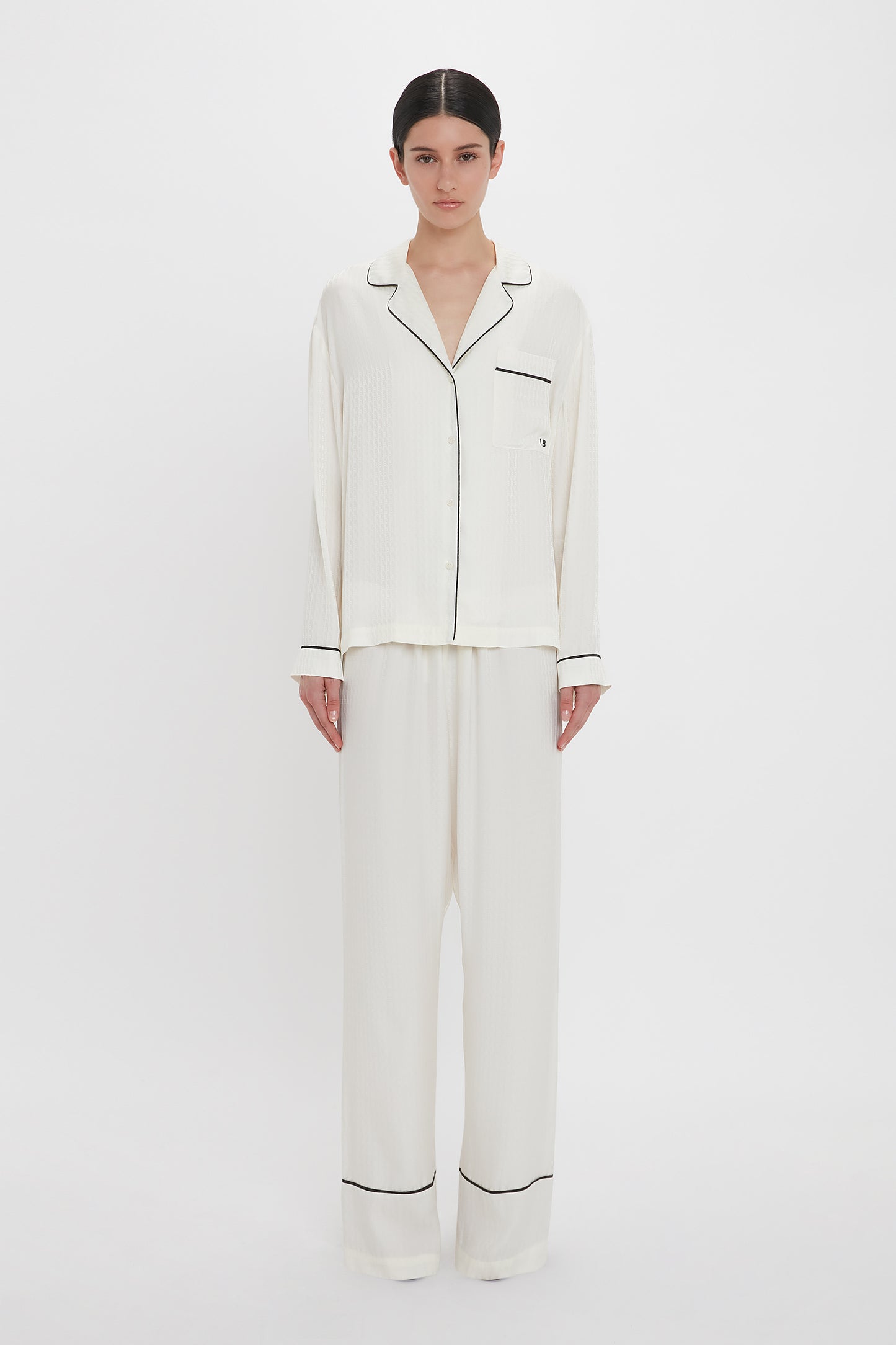 A person with dark hair stands against a white background wearing the Victoria Beckham VB Monogram Pyjama Sleep Set In Ivory.