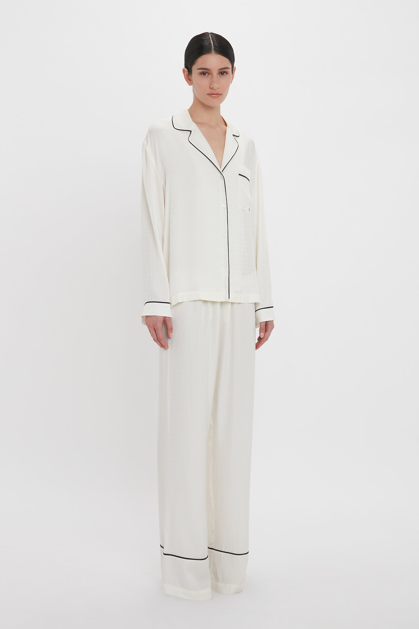 A person stands wearing a Victoria Beckham VB Monogram Pyjama Sleep Set In Ivory. The loungewear comprises a long-sleeve button-up top and matching wide-leg pants. The background is plain white.