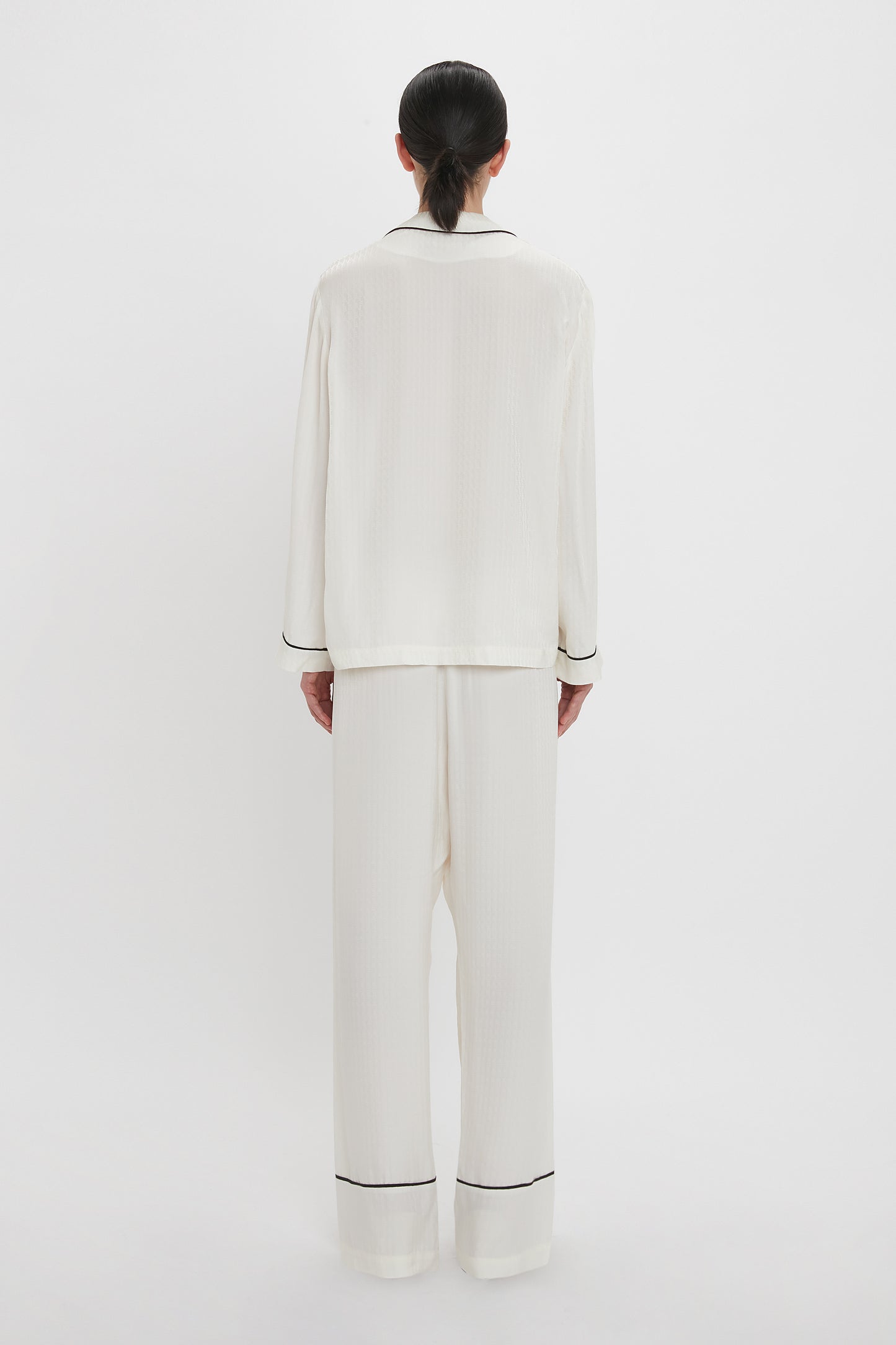 A person with dark hair is standing facing away, wearing a Victoria Beckham VB Monogram Pyjama Sleep Set in Ivory with black trim, against a plain white background.