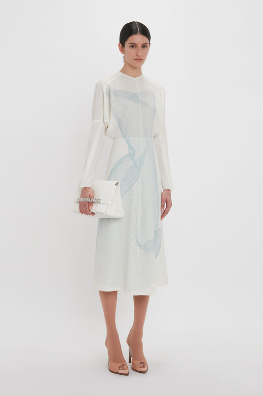 A woman in an elegant Victoria Beckham Long Sleeve Dolman Midi Dress In White-Blue Contorted Net, holding a white clutch, against a plain background.