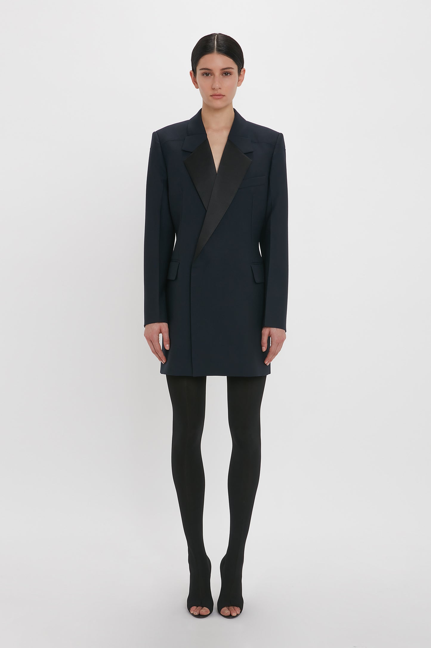 Person in an Exclusive Fold Shoulder Detail Dress In Midnight by Victoria Beckham, worn over black tights and open-toed shoes, standing against a plain white background.
