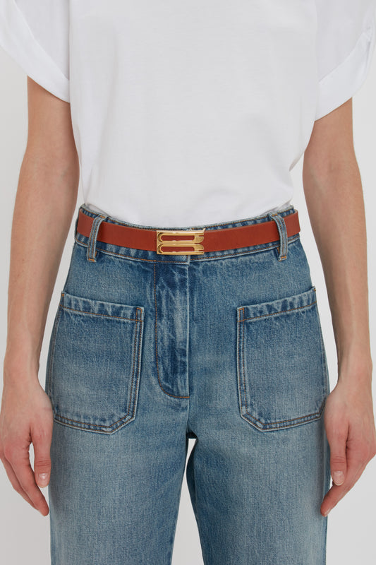 Close-up of a person wearing blue jeans and a white t-shirt, accented with a red and blue Victoria Beckham Exclusive Frame Buckle Belt In Tan Leather featuring gold hardware.