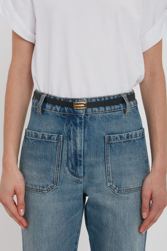 Close-up of a person wearing blue jeans with a Victoria Beckham Exclusive Micro Frame belt in black leather and a white t-shirt, focusing on the back pockets and belt area.