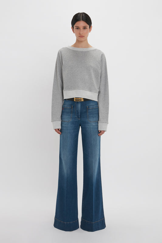 Woman standing in a studio, wearing a Victoria Beckham grey marl cropped sweatshirt and blue flared jeans with a thin belt.