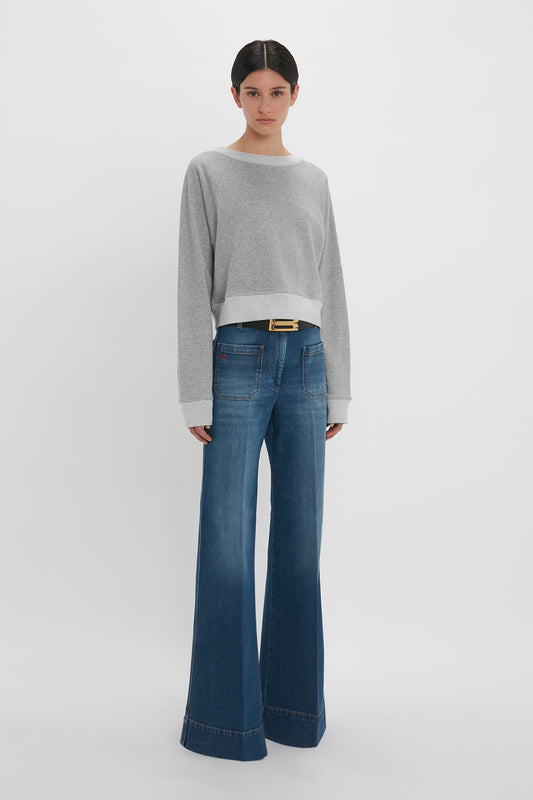 A woman stands against a white background, wearing a Victoria Beckham grey marl cropped sweatshirt and wide-legged blue jeans with a slim black belt.