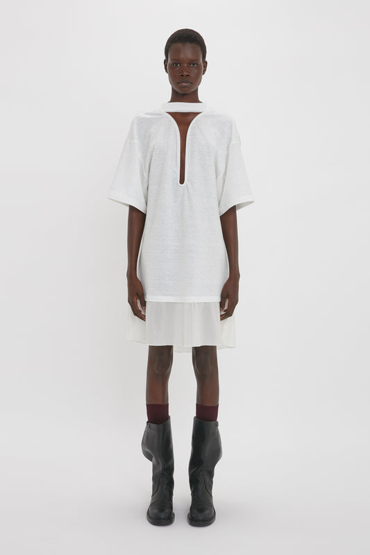 A black model wearing a Victoria Beckham white Frame Cut-Out T-Shirt Dress with a keyhole neckline and brown knee-high boots, standing against a plain background.