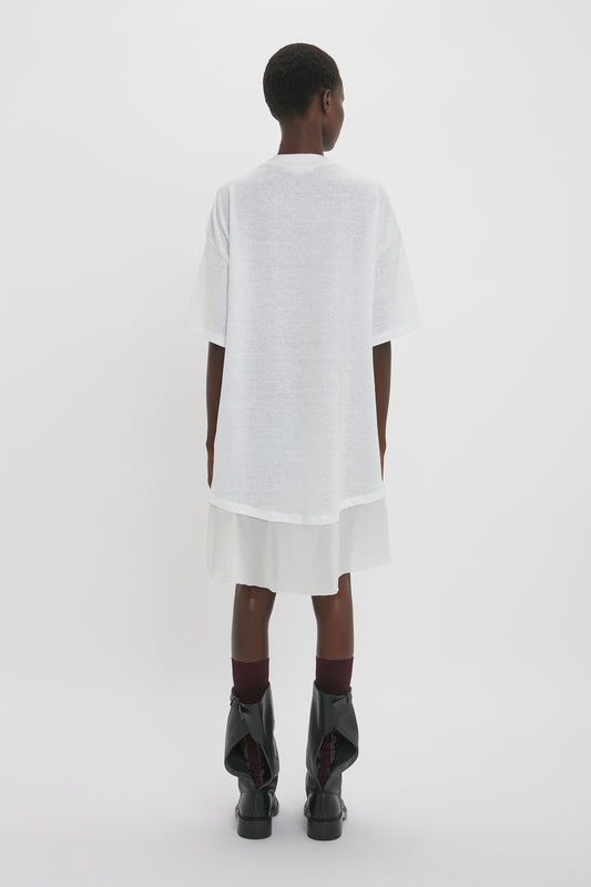 Rear view of a person standing in a Victoria Beckham Frame Cut-Out T-Shirt Dress In White and skirt, paired with black boots and maroon socks, against a plain background.