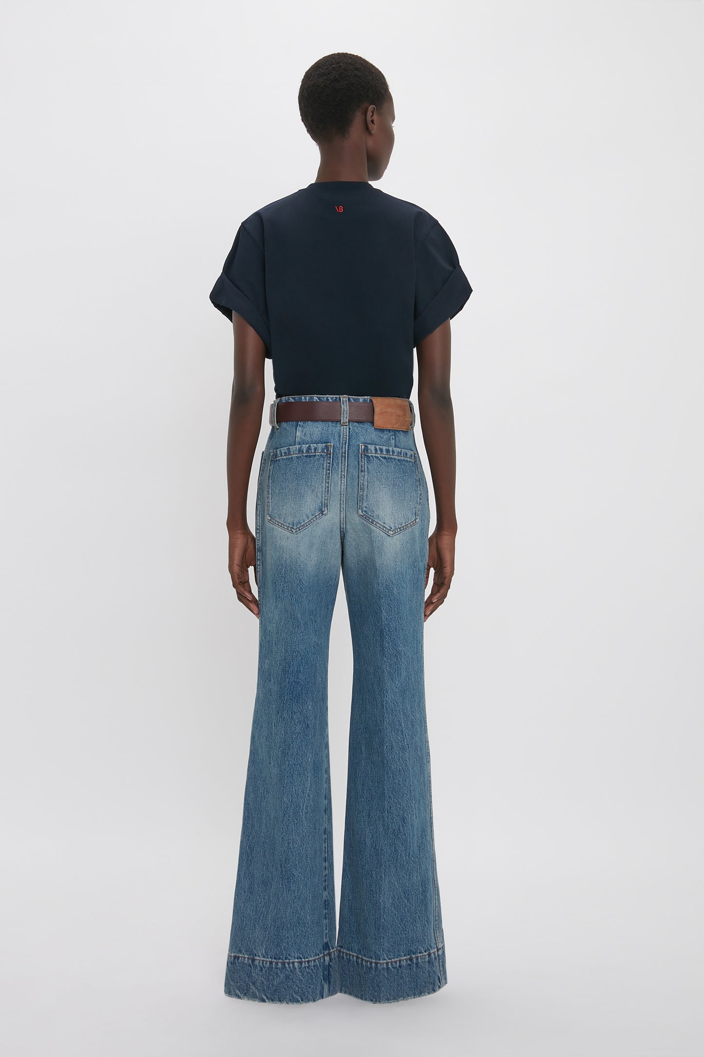 A person stands facing away, wearing an oversized Victoria Beckham Asymmetric Relaxed Fit T-Shirt In Navy with asymmetric folded sleeves, paired with high-waisted, wide-leg blue denim jeans featuring visible back pockets.