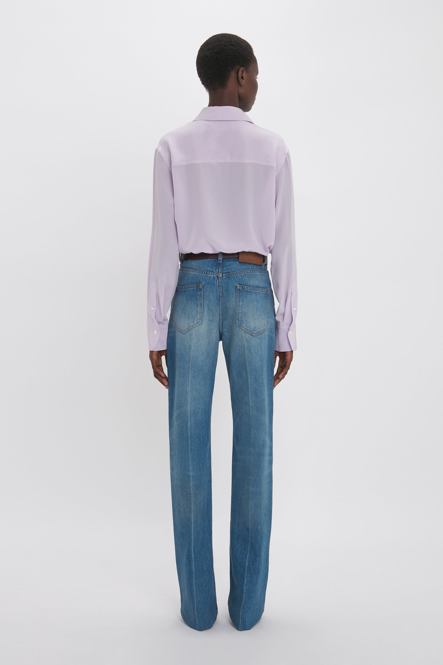 A person stands with their back to the camera, wearing a Victoria Beckham Asymmetric Ruffle Blouse in Petunia and classic blue jeans, against a plain white background.