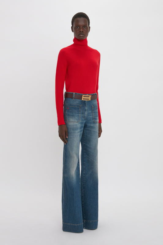 A woman wearing a red turtleneck and Alina Jean in Heavy Vintage Indigo Wash by Victoria Beckham with a gold-buckled belt stands against a white background.