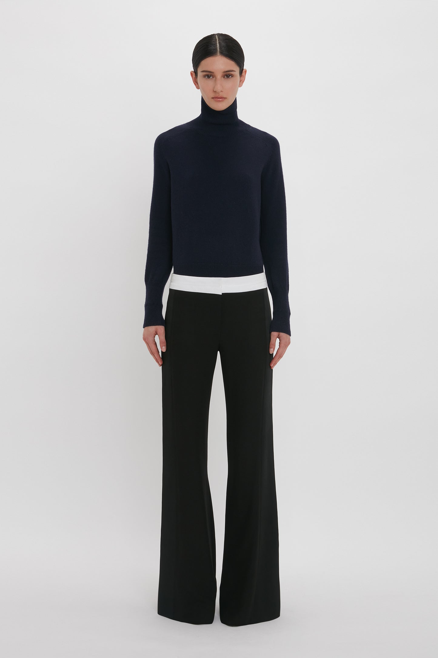 A person stands against a plain white background, wearing a dark navy turtleneck sweater, white undershirt, and the Side Panel Trouser In Black by Victoria Beckham with a wide-leg design. They have dark hair tied back.