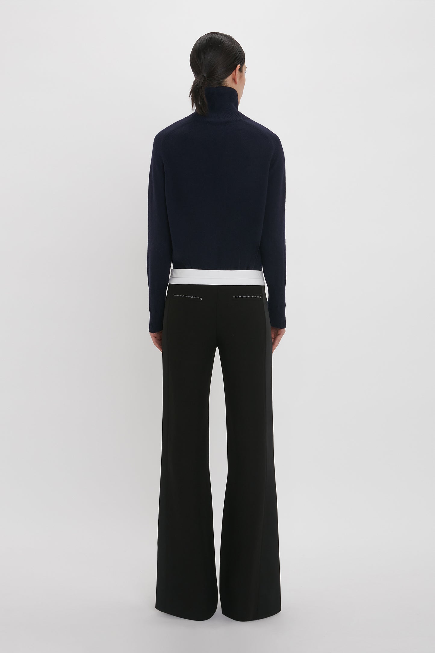 A person with dark hair is standing with their back to the camera, wearing a black turtleneck sweater, Victoria Beckham Side Panel Trouser In Black with a wide-leg design, and a white top underneath.