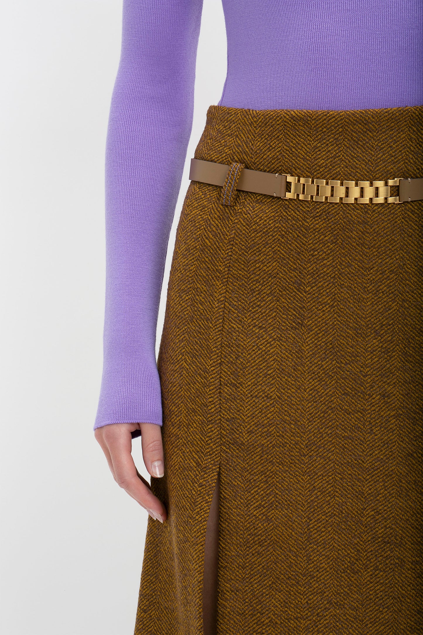 Close-up of a woman wearing a purple knitted polo neck jumper and a brown herringbone skirt with a Victoria Beckham UK Watch Strap Detail Belt in Khaki-Brown.