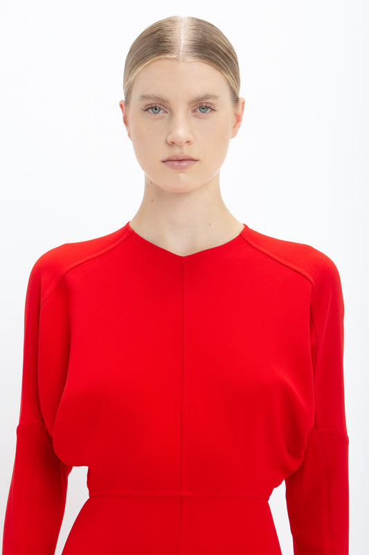 A woman with blonde hair styled back, wearing a vibrant red Victoria Beckham Dolman Midi Dress with a simple round neckline, standing against a white background.