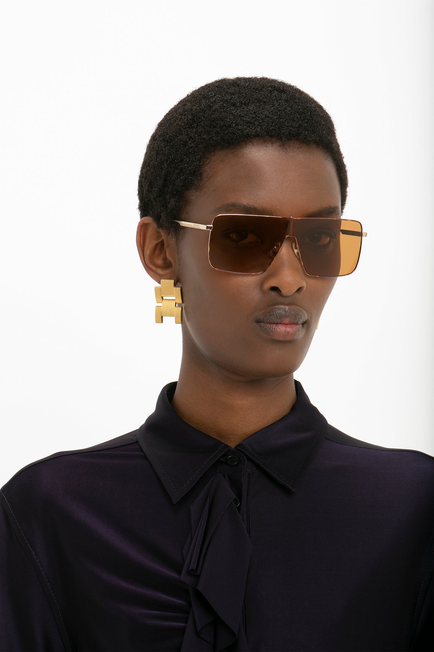 Person with short hair wearing Victoria Beckham Rectangular Visor Gold-Framed Sunglasses, gold geometric earrings, and a dark collared shirt.