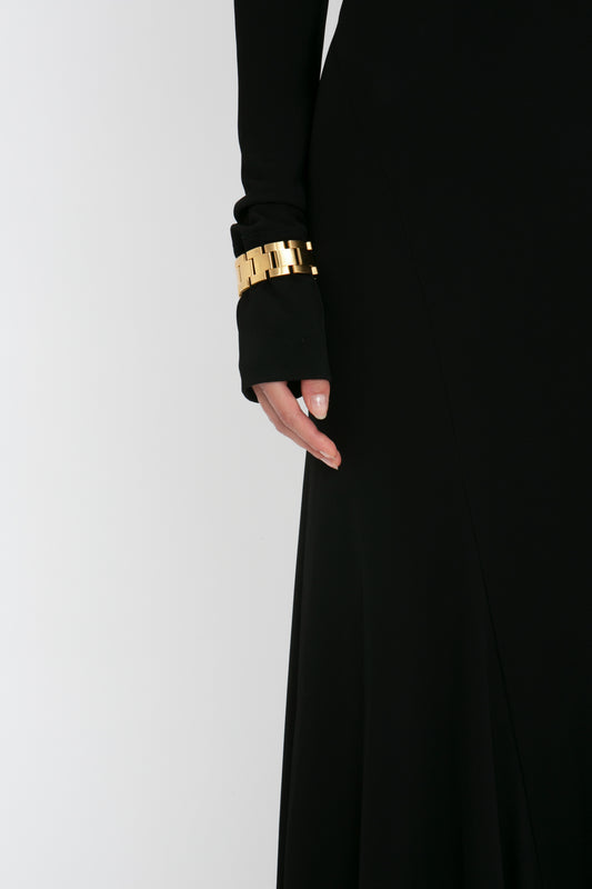 Close-up of a woman's arm in a Victoria Beckham Tie Detail Floor-Length Dress in Black gown with a gold bracelet, against a white background.