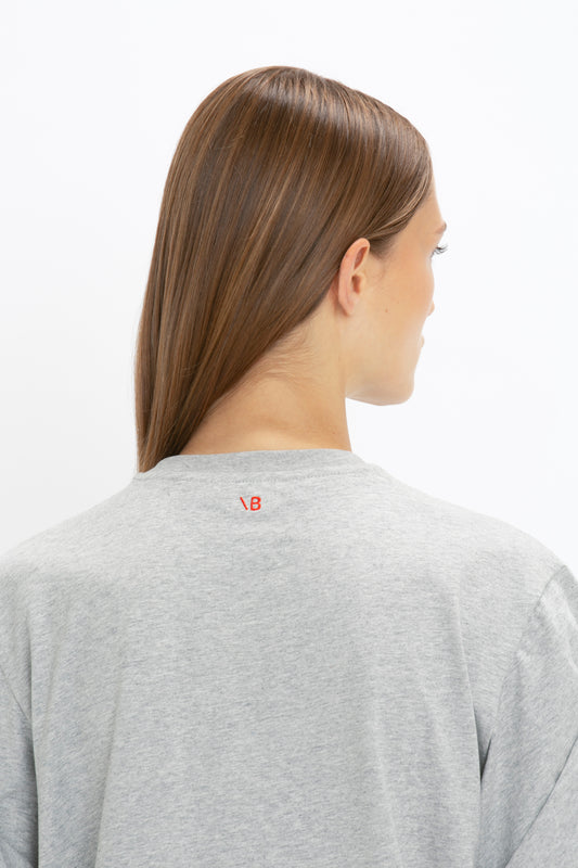 Woman in a Victoria Beckham Asymmetric Relaxed Fit T-shirt in Grey Marl with a small red logo on the back, viewed from behind with her brown hair styled straight.