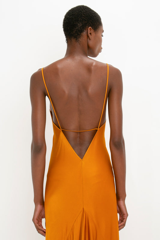 Rear view of a woman in an elegant Victoria Beckham floor-length camisole-slip dress in ginger, with a low-cut back and thin straps, standing against a white background.