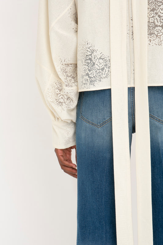 Close-up of a person wearing denim jeans and a white lace-trimmed shawl with blouson sleeves, focusing on the Victoria Beckham Asymmetric Gather Detail Top In Cream from mid-thigh to wrist.