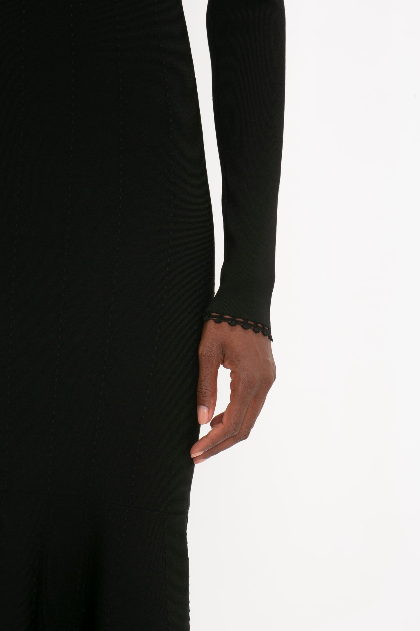 Close-up of a person wearing a Victoria Beckham VB Body Long Sleeve V Neck Dress In Black with a scalloped edge on the sleeve, showcasing their hand with well-manicured nails against a white background.